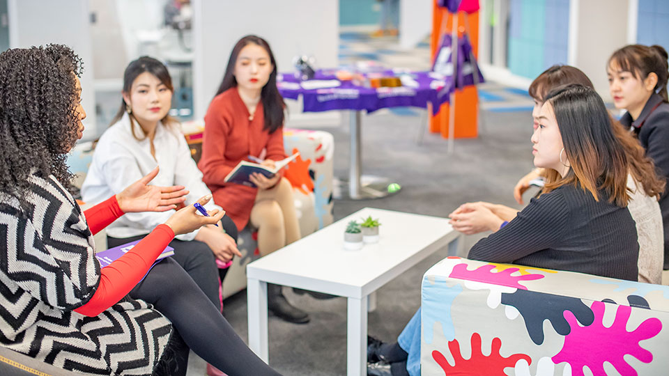 A group of people sit in a modern office setting, engaged in discussion around a white table, with colourful furniture in the background.